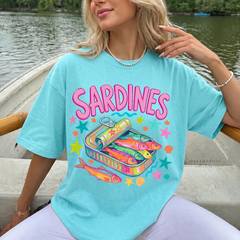 Comfort Colors 1717 lagoon blue t-shirt with the word "Sardines" in pink, above a colorful illustration of sardines in a can. The graphic includes stars, shells, and fish in vibrant colors. The shirt has short sleeves.