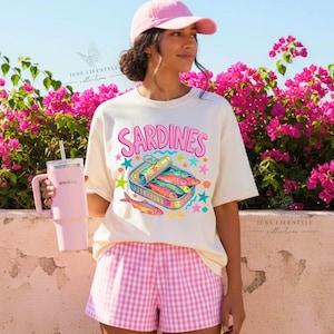 A woman wearing a ivory-colored t-shirt with the word "Sardines" and a colorful sardine can graphic. She is wearing a pink baseball cap and pink and white checkered shorts. She is holding a pink tumbler.