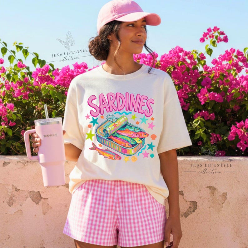 A woman wearing a ivory-colored t-shirt with the word "Sardines" and a colorful sardine can graphic. She is wearing a pink baseball cap and pink and white checkered shorts. She is holding a pink tumbler.