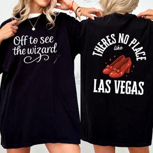 May include: Black t-shirt with white text that reads "Off to see the wizard" on the front. The back of the shirt has the text "There's no place like Las Vegas" with an illustration of red high-heeled shoes.