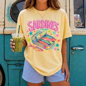 Yellow t-shirt with the word "SARDINES" in pink letters above a colorful sardine can illustration. The can contains several brightly colored fish. The shirt is paired with blue and white striped shorts.