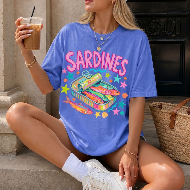 Comfort Colors 1717 Flo- blue t-shirt with the word "Sardines" in pink, above a colorful illustration of sardines in a can. The graphic includes stars, shells, and fish in vibrant colors. The shirt has short sleeves.