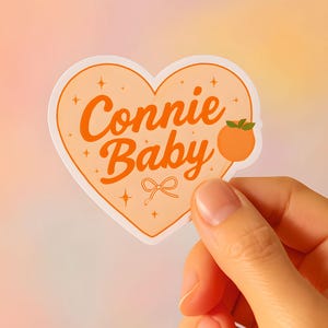 May include: A heart-shaped sticker with the words "Connie Baby" in orange script. The sticker features a peach illustration and a small bow. The background is a gradient of peach and yellow.
