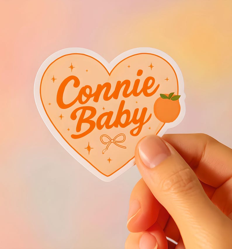 May include: A heart-shaped sticker with the words "Connie Baby" in orange script. The sticker features a peach illustration and a small bow. The background is a gradient of peach and yellow.