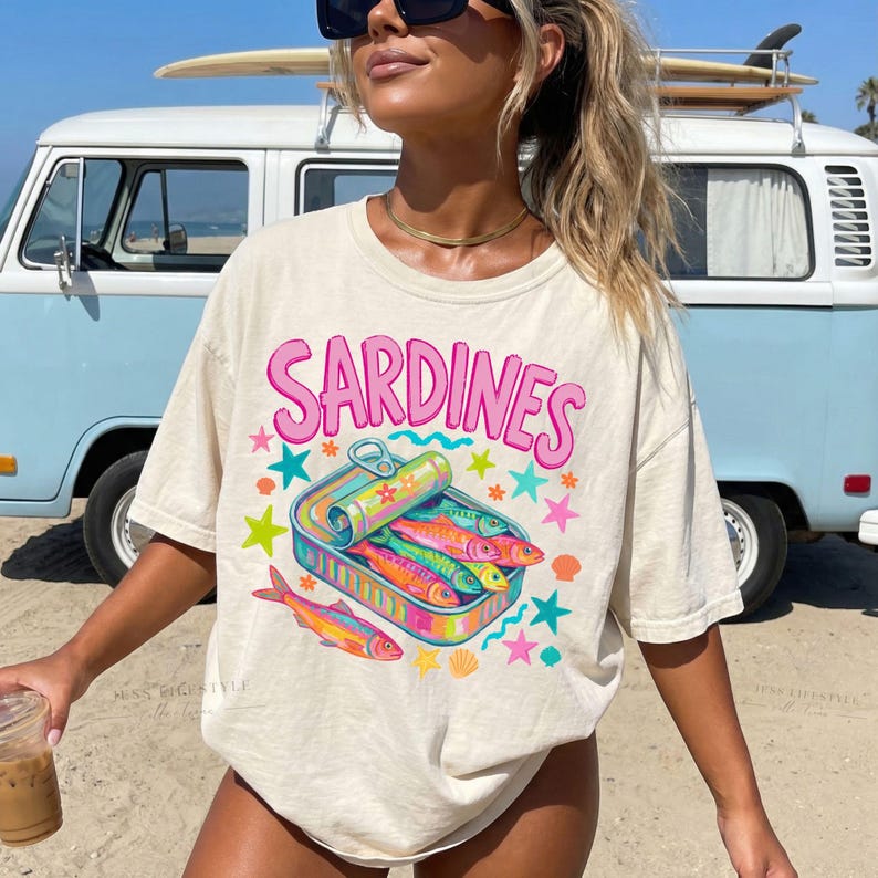 Beige t-shirt with the word "SARDINES" in pink bubble letters. The shirt features a colorful graphic of sardines in a can, surrounded by stars and seashells. A casual, beach-themed tee.