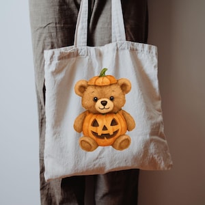 May include: A cream tote bag with a watercolour illustration of a teddy bear in a pumpkin costume. The bear is brown with a pumpkin hat and a carved pumpkin body. The bag has a simple design with two handles.