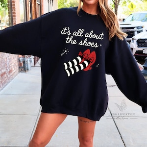 Puede incluir: Sudadera negra con la frase "it's all about the shoes" y un gráfico de zapatos rojos con lazos y calcetines a rayas blancas y negras. La sudadera tiene un corte holgado y mangas largas.