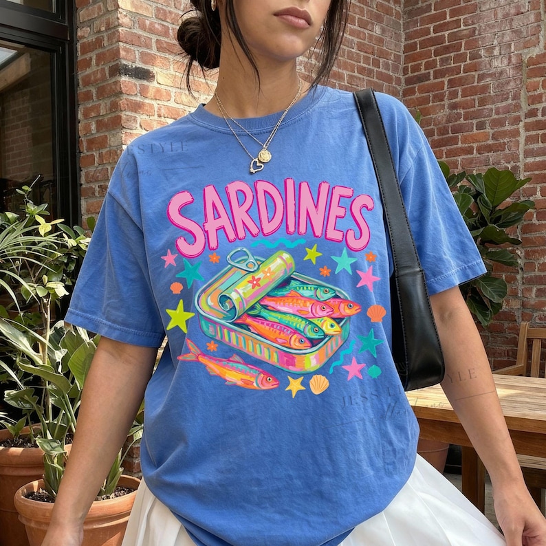 Comfort Colors 1717 Flo- blue t-shirt with the word "Sardines" in pink, above a colorful illustration of sardines in a can. The graphic includes stars, shells, and fish in vibrant colors. The shirt has short sleeves.