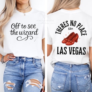 May include: White t-shirt with the text "Off to see the wizard" on the front and "There's no place like Las Vegas" with ruby red shoes on the back. The shirt is paired with blue jeans.