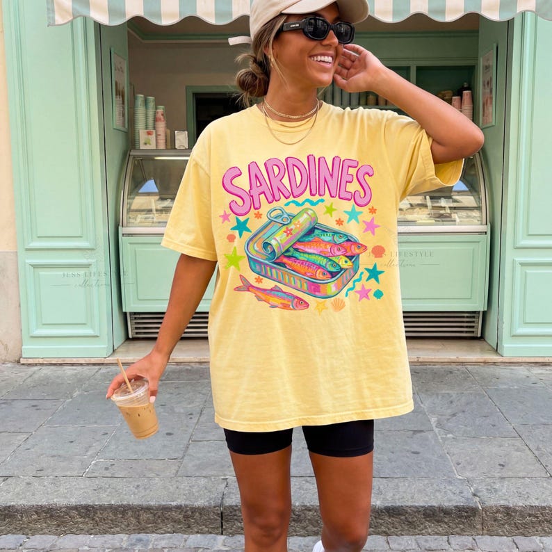 Butter Yellow t-shirt with the word "SARDINES" in pink letters above a colorful sardine can graphic. The graphic includes sardines, stars, and a rainbow design. The shirt is worn by a person wearing black shorts and sunglasses.