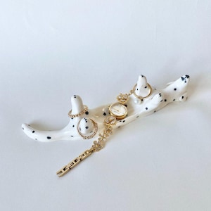 Op de afbeelding: Een witte keramische sieradenhouder met zwarte stippen, met gouden ringen en een gouden horloge met een bloemenontwerp. De houder heeft de vorm van een lange, gebogen vorm.