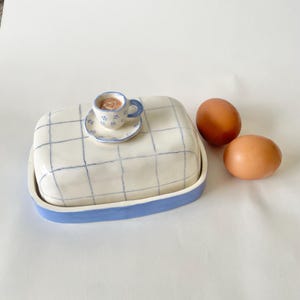Può includere: Un portaburro in ceramica con base bianca caratterizzata da un motivo a griglia blu e un bordo blu solido. Il coperchio è sormontato da una tazzina con piattino in miniatura decorati con piccoli fiori blu e un cuoricino, accanto a due uova marroni.