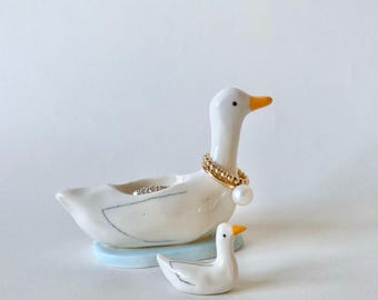 Puede incluir: Un joyero con forma de ganso de cerámica blanca con un anillo de oro y un collar de perlas. Una figura de ganso más pequeña está a su lado. La base es azul claro y el ganso tiene un pico naranja.