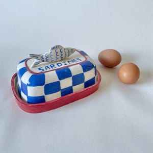 Op de afbeelding: Een keramische botervloot met een blauw-wit geblokt patroon en een rode onderkant. Het deksel heeft een handvat in de vorm van een gevormde vis en de tekst SARDINES in blauwe letters. Twee bruine eieren liggen naast de botervloot.