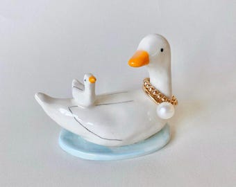 Puede incluir: Figura de pato de cerámica blanca con pico naranja y un patito en la espalda. El pato lleva un collar dorado con colgante de perla y está sobre una base azul claro. Artículo decorativo.