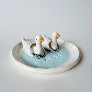 May include: A white ceramic ring dish featuring two white duck figurines with orange beaks. The dish has a light blue centre with the embossed text m & k and a black grid pattern on the rim. Two metal rings are placed around the necks of the ducks.