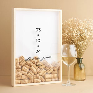 May include: A wooden box with a glass front filled with wine corks. The box has a white background with the numbers 03, 10, and 24 written in black. The text "alice + simon wedding" is written in black cursive script. A glass of white wine sits on a table next to the box.