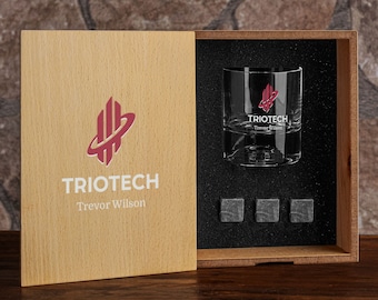 May include: A light-colored wooden box containing a clear glass tumbler and three square gray stone cubes. The box lid and glass feature a red logo with the text TRIOTECH and the name Trevor Wilson printed in white.