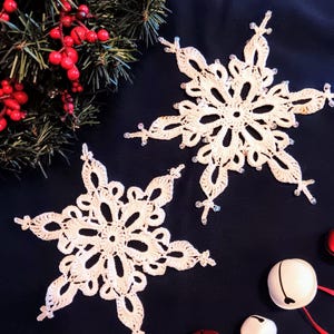 May include: Two white crocheted snowflakes with beaded edges on a dark blue background. There are also three jingle bells, two white and one red, with red ribbon.