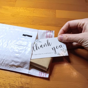 May include: A white padded envelope with a small brown box inside. A white card with the text "thank you for supporting my small business" is being pulled out of the envelope.