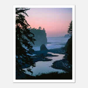 May include: A framed print of a coastal landscape at dusk. The scene features a calm body of water leading to the ocean, with a rocky cliff and trees silhouetted against a pink and purple sky. The print is in a white frame.