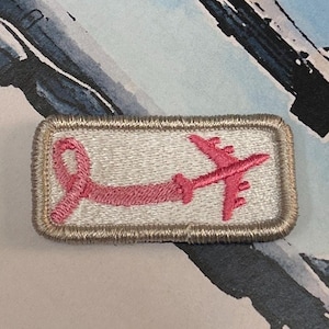 May include: Embroidered patch with a pink aeroplane linked to a pink ribbon, on a light grey background. The patch is rectangular with a tan border.