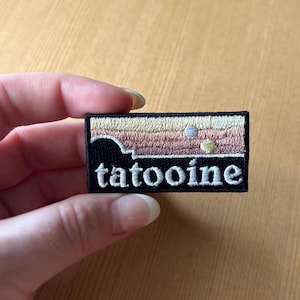Peut inclure: Écusson brodé avec le mot "tatooine" en blanc, présentant un paysage dans des tons de rose, beige et noir. L'écusson est rectangulaire et a une bordure noire.