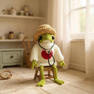May include: A green crocheted frog doll wearing a tan straw-style hat and a white knitted sweater with a red heart applique on the chest. The frog has black bead eyes and a black cord necklace with a heart-shaped charm, sitting on a small stool.