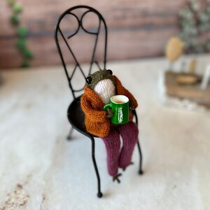 May include: A knitted frog wearing a brown sweater and purple trousers sits on a black metal chair, holding a green mug with brown liquid and the text "Nescafé" in white.