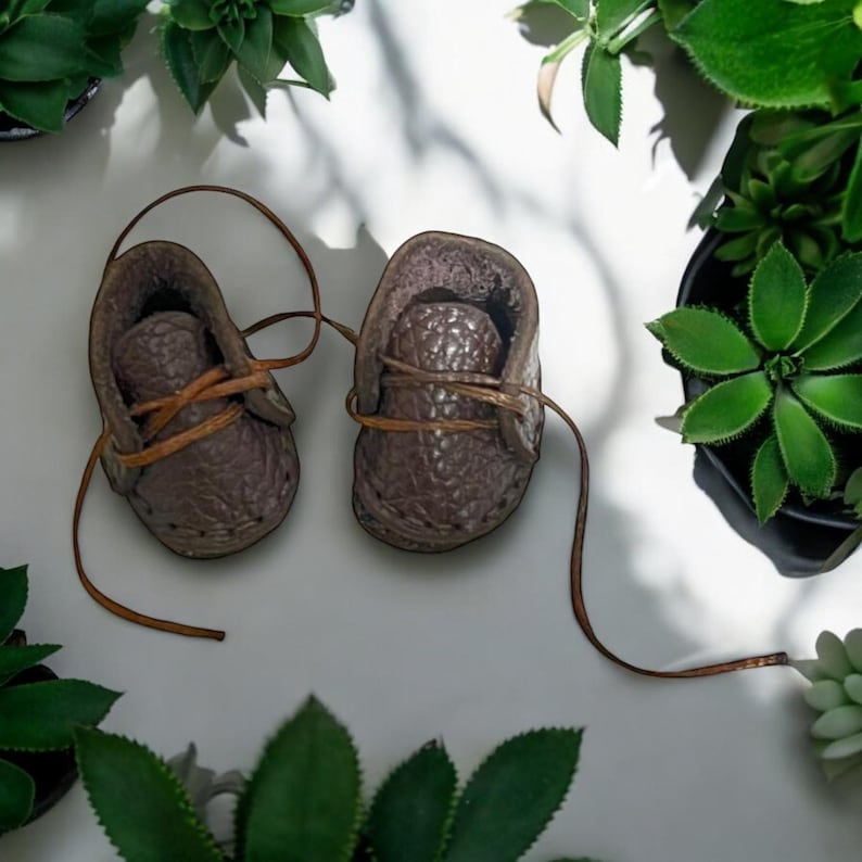 May include: A pair of brown leather baby shoes with laces. The shoes are on a white surface with green plants in the background.