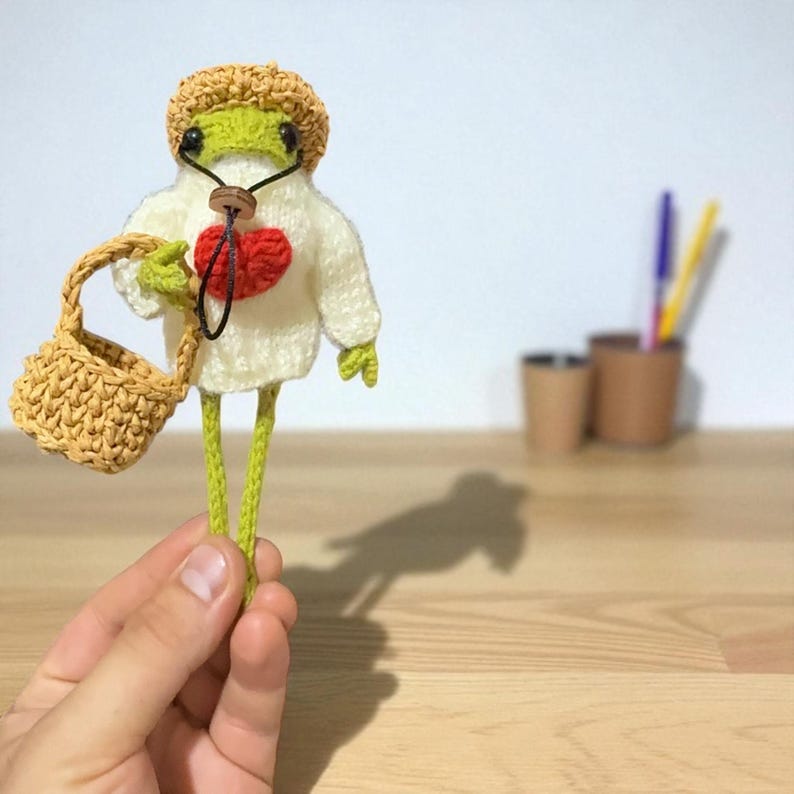 May include: A crocheted green frog figurine wearing a tan hat and a white sweater with a red heart appliqu&eacute;. The frog has long thin legs and holds a small tan crocheted basket. A thin black cord with a small wooden bead hangs around its neck.