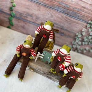 May include: Four knitted frog figurines wearing brown overalls and red and white striped jumpers. Each frog is holding a red mushroom. The frogs are sitting on a wooden surface with a small green house and a sign that reads "Drink World".