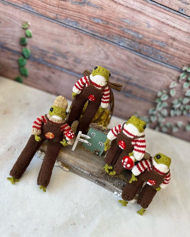 May include: Four knitted frog figurines wearing brown overalls and red and white striped jumpers. Each frog is holding a red mushroom. The frogs are sitting on a wooden surface with a small green house and a sign that reads "Drink World".