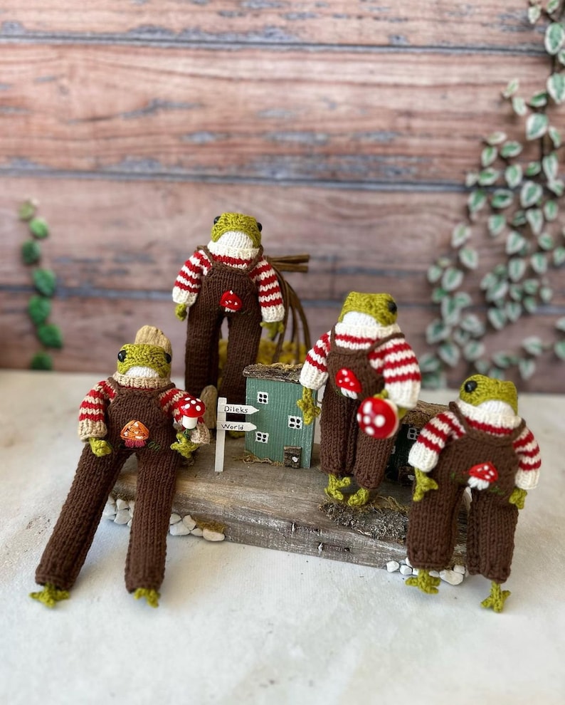 May include: Four knitted frog figurines wearing brown overalls and red and white striped jumpers. The frogs are holding red and white mushrooms. The frogs are sitting on a wooden surface with a small wooden house and a sign that says "Dileks World".