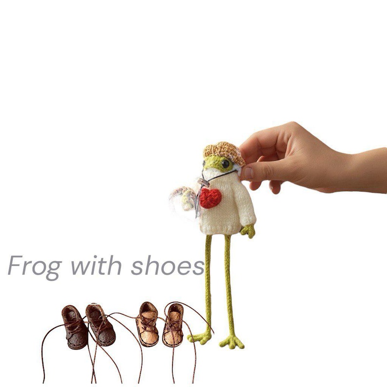 May include: A small green crocheted frog figurine wearing a white sweater, a tan hat, and a red heart accessory, positioned above four miniature brown leather-style shoes with laces. The text "Frog with shoes" is printed in grey on the left side.