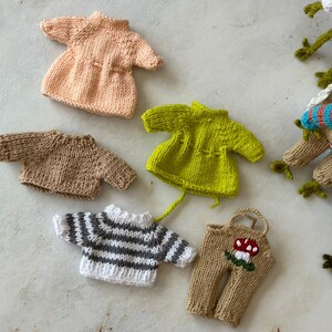 Puede incluir: Una colección de ropa de muñeca tejida, que incluye un vestido rosa, un vestido verde, un suéter marrón, un suéter a rayas y un mono marrón con un diseño de hongo.