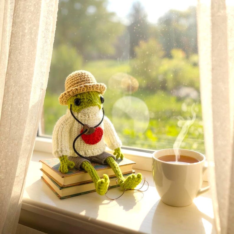 May include: A crocheted green frog doll wearing a beige sun hat, a white sweater with a red heart, and grey shorts, sitting on a stack of two books next to a white ceramic mug filled with a steaming hot beverage near a window.