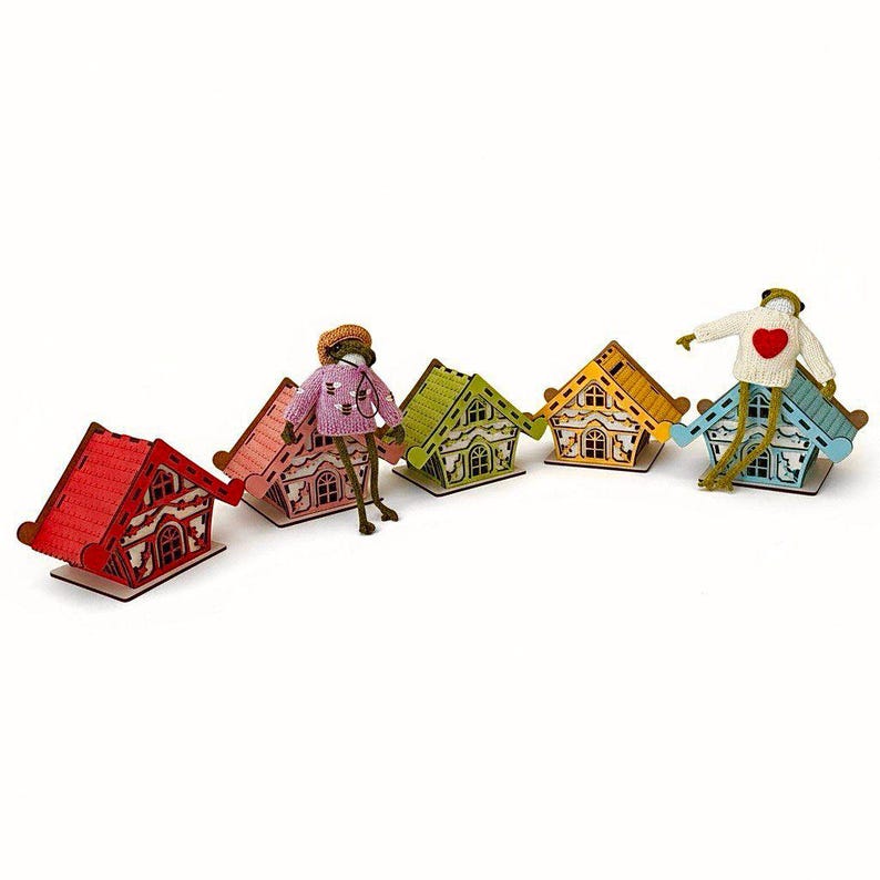 May include: A set of five small wooden houses in red, pink, light green, yellow, and light blue, each with intricate cut-out details. Two frog figurines are included: one wearing a pink bee-patterned sweater and one in a white sweater with a red heart.