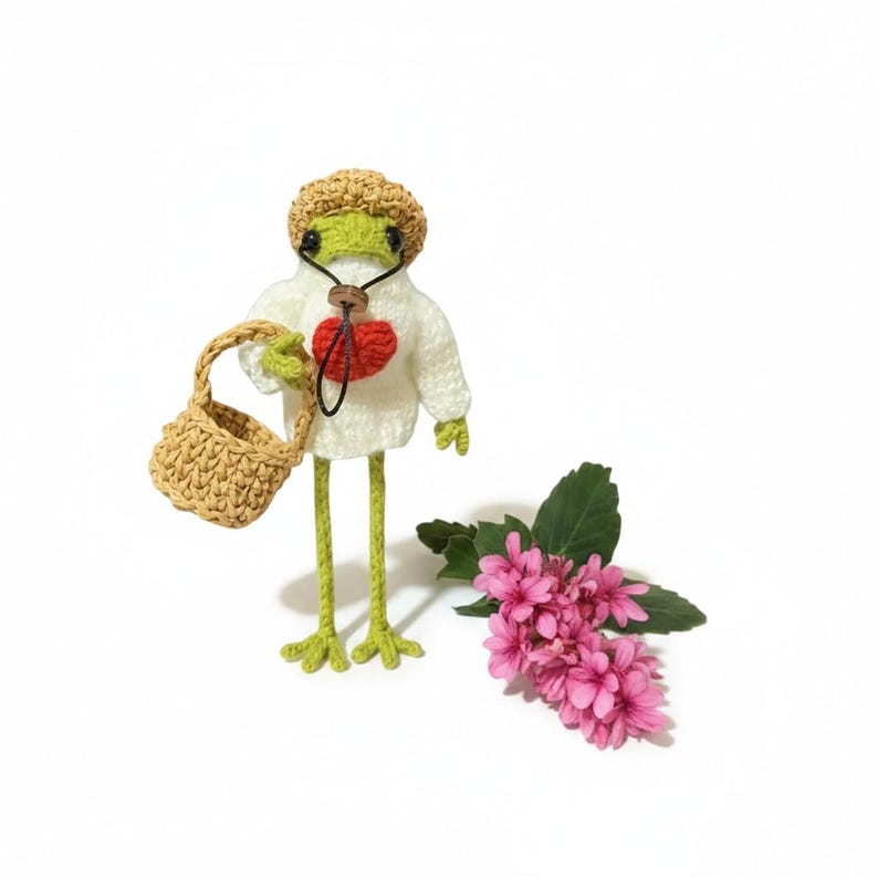 May include: A crocheted green frog doll wearing a tan straw-style hat and a white sweater with a red heart detail. The frog holds a small tan woven basket in one hand and stands next to a cluster of pink flowers with green leaves on a white surface.
