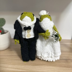 Op de afbeelding: Een gebreid kikkerpaar gekleed in trouwtenue. De bruidegom draagt ​​een zwart smoking met een wit overhemd en een strikje. De bruid draagt ​​een witte jurk met een witte sluier.
