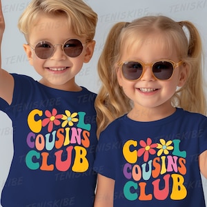 K&ouml;nnte beinhalten: Zwei Kinder tragen marineblaue T-Shirts mit einem bunten "Cool Cousins Club"-Motiv. Die Shirts haben eine Retro-Schriftart und Blumenakzente. Die Kinder l&auml;cheln und zeigen den Peace-Zeichen.