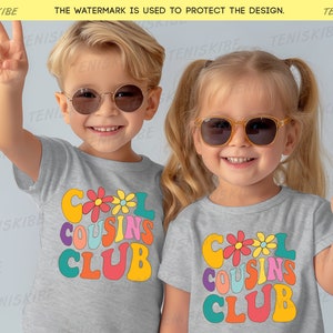 K&ouml;nnte beinhalten: Zwei Kinder tragen graue T-Shirts mit dem Text "Cool Cousins Club" in bunter Retro-Schrift. Die Kinder tragen Sonnenbrillen und l&auml;cheln.