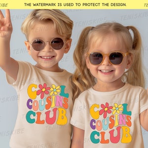K&ouml;nnte beinhalten: Zwei Kinder tragen wei&szlig;e T-Shirts mit dem Text "Cool Cousins Club" in bunter Retro-Schrift und Blumen. Die Kinder tragen Sonnenbrillen und l&auml;cheln.