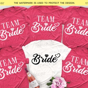 Puede incluir: Cinco camisetas rosas y blancas con el texto "Team Bride" en letras blancas con un gráfico de diamante y un corazón. Las camisetas están dispuestas en círculo con una botella de vino y flores en el centro.