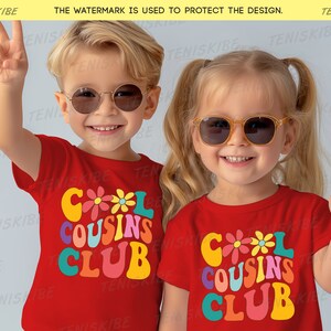K&ouml;nnte beinhalten: Zwei Kinder tragen rote T-Shirts mit dem Text "Cool Cousins Club" in bunter Retro-Schrift. Die Shirts haben ein Blumendesign. Die Kinder tragen Sonnenbrillen und l&auml;cheln.