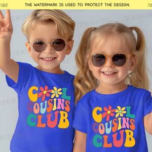 K&ouml;nnte beinhalten: Zwei Kinder tragen blaue T-Shirts mit dem Text "Cool Cousins Club" in bunter Retro-Schrift und Blumen. Die Kinder tragen Sonnenbrillen und l&auml;cheln.