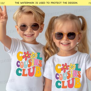K&ouml;nnte beinhalten: Zwei Kinder tragen wei&szlig;e T-Shirts mit einem bunten "Cool Cousins Club"-Motiv. Das Motiv zeigt eine Retro-Schriftart und Blumen. Die Kinder l&auml;cheln und machen Peace-Zeichen.