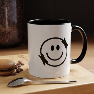 Könnte beinhalten: Weiße Keramiktasse mit schwarzem Rand und Henkel. Die Tasse hat ein schwarz-weißes Smiley-Gesicht-Design.