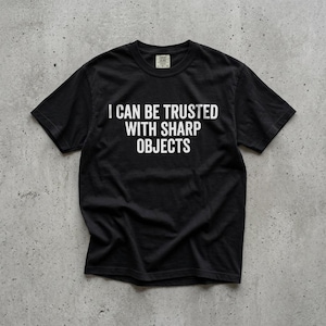 May include: A black short-sleeve t-shirt featuring the white text I CAN BE TRUSTED WITH SHARP OBJECTS printed in a bold, sans-serif font across the chest. The garment has a crew neckline and a standard fit, showcasing a clean, graphic design.