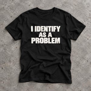 May include: A black short-sleeved t-shirt featuring the white text I IDENTIFY AS A PROBLEM printed in a bold, sans-serif font across the centre of the chest. The garment has a crew neckline and a standard fit, displaying a simple, clean design.